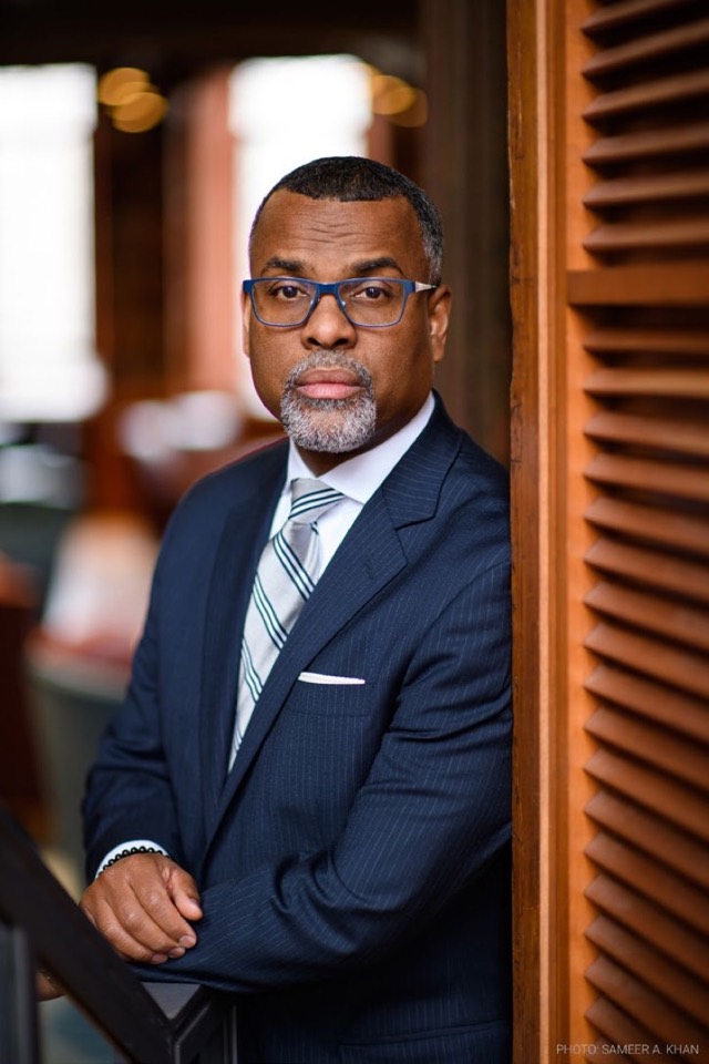 Dr. Eddie Glaude Jr. in a navy pinstripe suit, standing beside wooden shutters