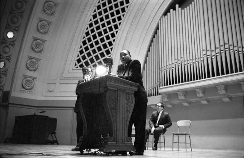 Black and white photo of Dr. Martin Luther King Jr. standing at a carved wooden podium with microphones on the stage of Hill Auditorium, with organ pipes and an ornate arch behind him and a man in a suit seated at right.