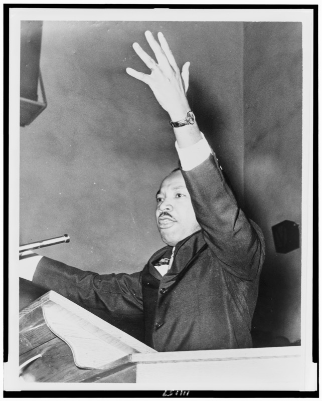 Dr. Martin Luther King Jr. at a wooden pulpit beside a microphone, his left arm raised high as he speaks.