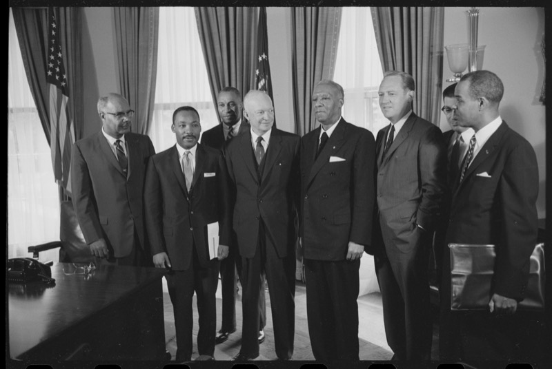 Eight men in dark suits stand in a row in a White House office with flags behind them. Dr. King, second from left, holds papers, and President Eisenhower stands at center.