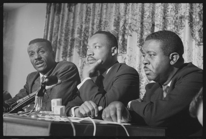 Rev. Fred Shuttlesworth speaks into a microphone at a press table while Dr. King, center, rests his chin on his hand and Rev. Ralph Abernathy listens, in front of a floral curtain.