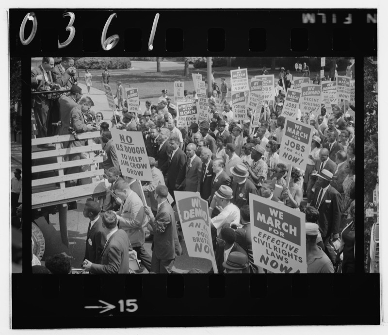 Civil rights leaders in suits march through a dense crowd holding signs that demand jobs, voting rights and effective civil rights laws, with Dr. King among them and press photographers on a truck at left.