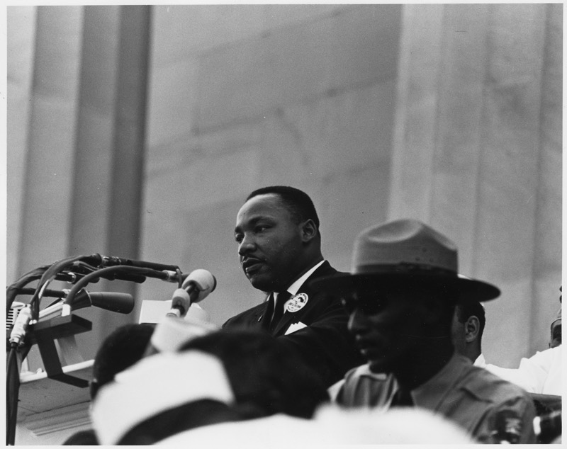 Black and white photograph of Dr. Martin Luther King Jr. speaking into a cluster of microphones, a March on Washington button on his lapel, with tall stone columns behind him and a park ranger in a campaign hat in the right foreground.