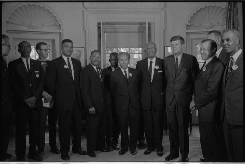 A row of civil rights leaders in suits and march buttons stands with President Kennedy in the Oval Office. Dr. King is fifth from left, and Kennedy stands right of center.