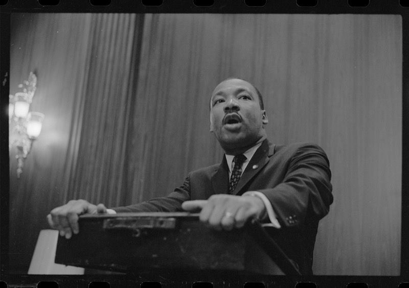 Black and white photograph of Dr. Martin Luther King Jr. leaning over a lectern with both hands on its edge, speaking, seen from a low angle against a draped wall with a lit sconce at left.