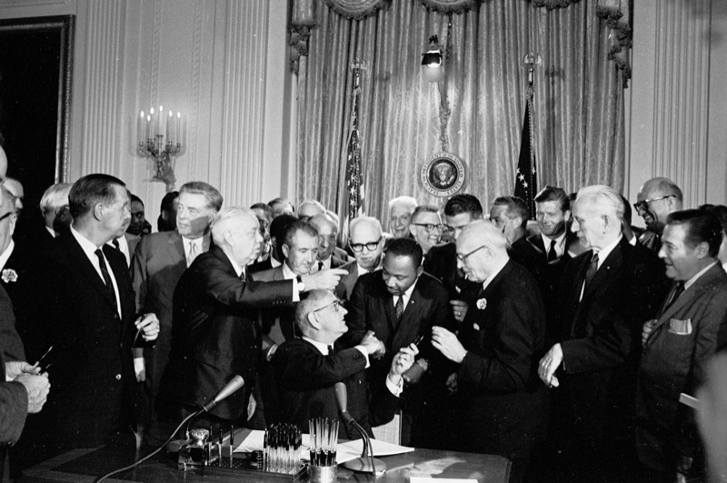 Black and white photograph in the White House East Room: President Lyndon B. Johnson, seated at a desk with microphones and a holder of pens, hands a pen to Dr. Martin Luther King Jr., who stands among a crowd of men in suits before curtains, flags and the presidential seal.