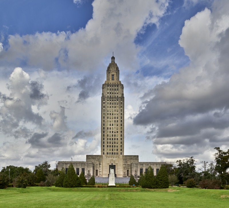 Color photograph of the tall Art Deco tower of the Louisiana State Capitol in Baton Rouge under billowing clouds, seen across a green lawn with a white memorial statue in front of the entrance.