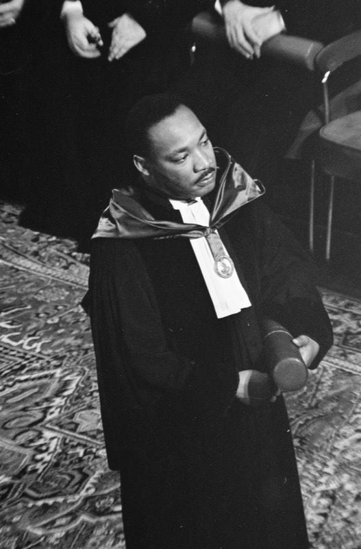 Black and white photo of Dr. Martin Luther King Jr. in an academic gown with a satin hood, white bands and a medallion, holding a cylindrical diploma case and standing on a patterned carpet.