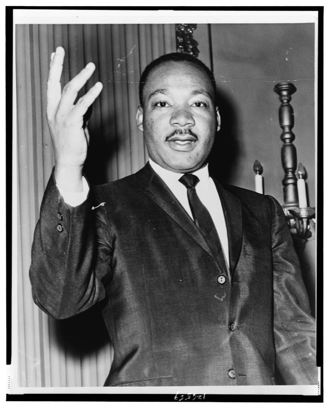 Black and white portrait of Dr. Martin Luther King Jr. facing the camera with a slight smile, his right hand raised beside his head, wearing a dark suit and tie, with a curtain and a candle sconce behind him.